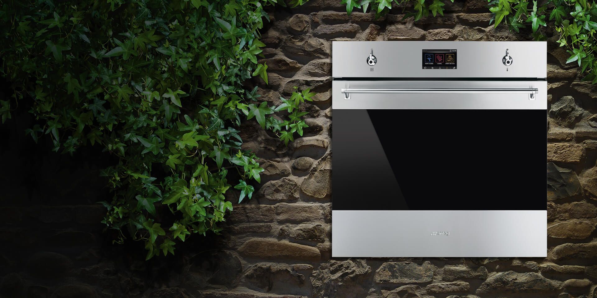 Classica built-in oven featuring a stainless steel and black glass design, photographed front view against a rustic stone wall with green foliage