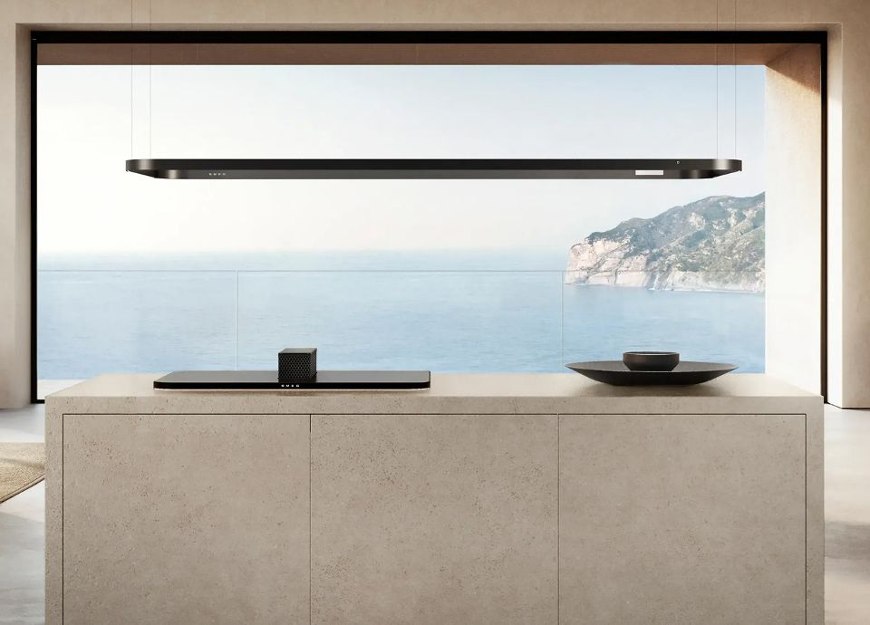 A sandy-beige stone kitchen island with a raised black hob and a black lighting rail, overlooking a blue ocean.