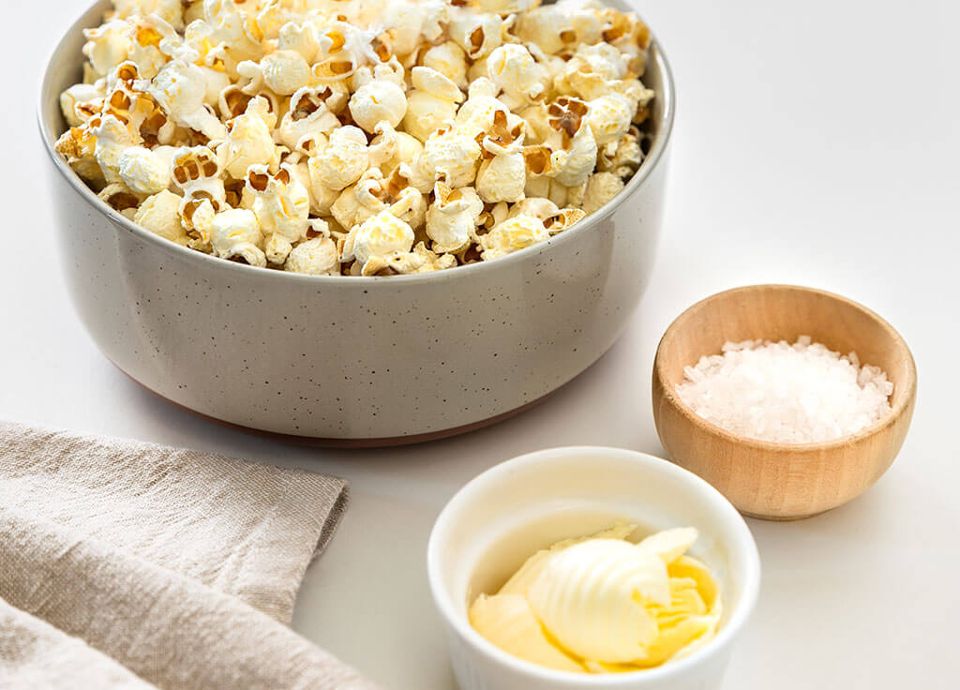 A large ceramic bowl of popcorn next to small dishes of swirled butter and sea salt on a bright white surface.