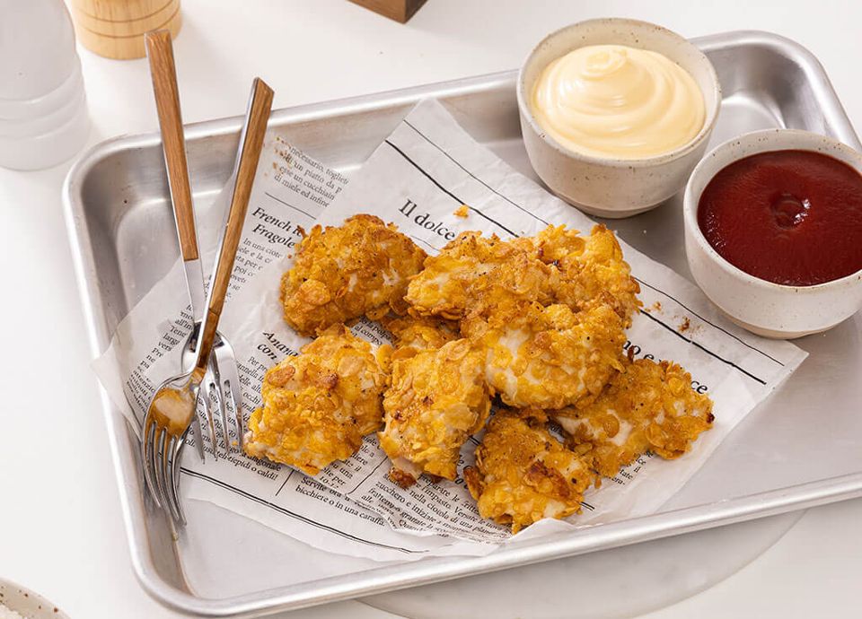 Golden crusted chicken on newsprint paper in a metal tray, served with cream and red sauces and wooden forks.