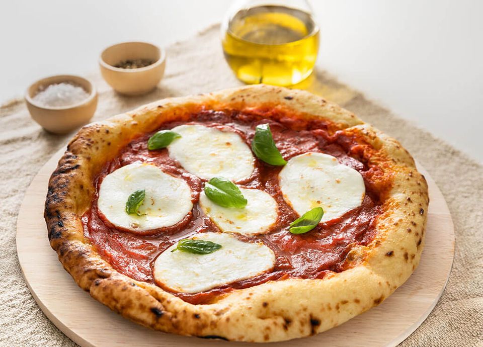 Fresh pizza with melted mozzarella rounds and basil on a ceramic stone, with olive oil and spices in the background.
