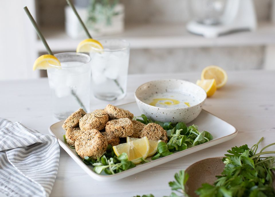 Crispy falafel patties on greens with a bowl of creamy dip, lemon wedges, and two iced drinks on a white table.