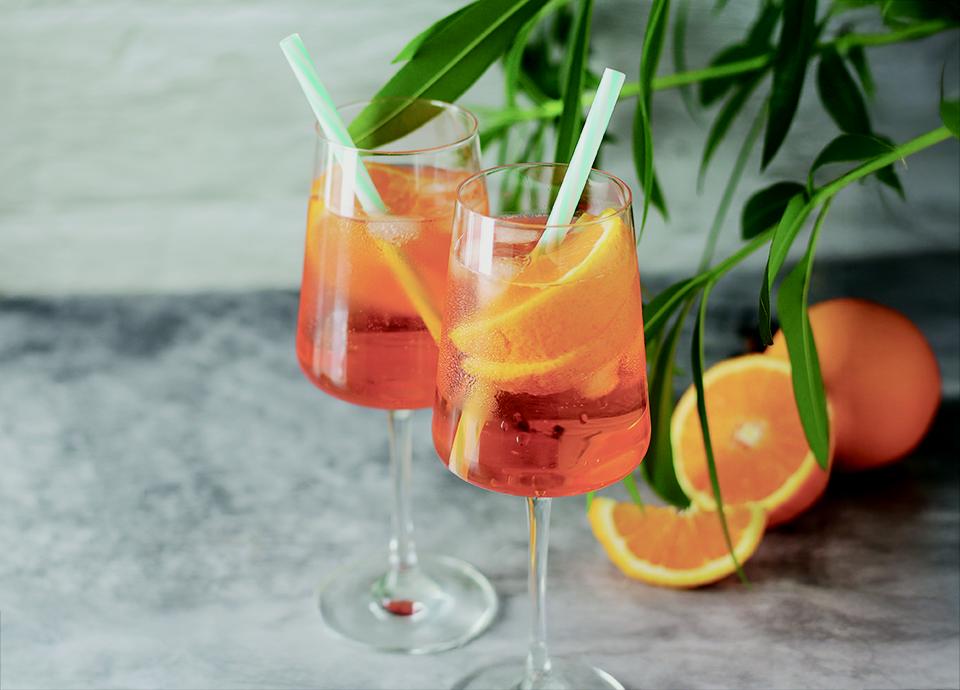 A pair of wine glasses filled with Orange Spritz, ice, straws, and orange slices, set against a textured background with green leaves.