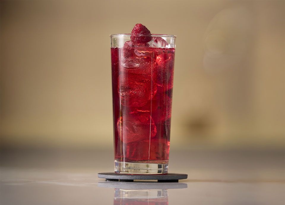 A bright red drink with ice and a raspberry in a tall glass, placed on a dark gray coaster.