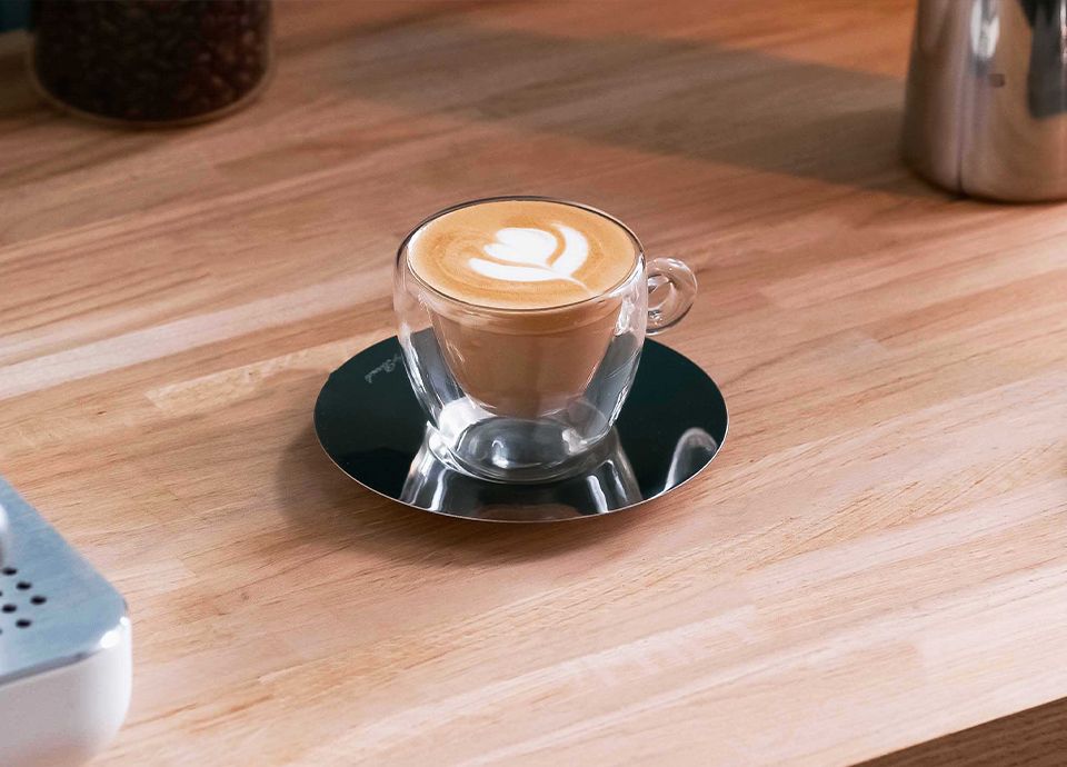 Cappuccino with latte art in a transparent glass cup on a dark reflective saucer, placed on a wooden table.