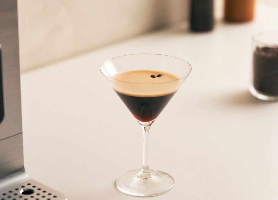 An Espresso Martini with thick foam and coffee beans in a crystal glass, next to a Smeg coffee machine.