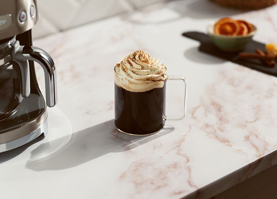 Ginger coffee in a glass cup topped with a generous dollop of whipped cream, served on a marbled countertop next to a Smeg coffee machine.
