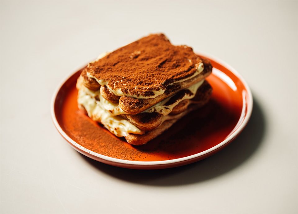 A square slice of tiramisu layered with mascarpone cream and coffee-soaked biscuits, dusted generously with cocoa powder and served on an orange plate.