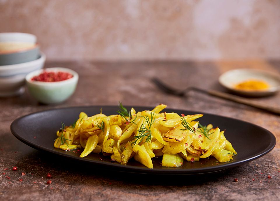 Yellow fennel with turmeric, garnished with dill, served on a black oval plate, with pink berries and small bowls in the background.