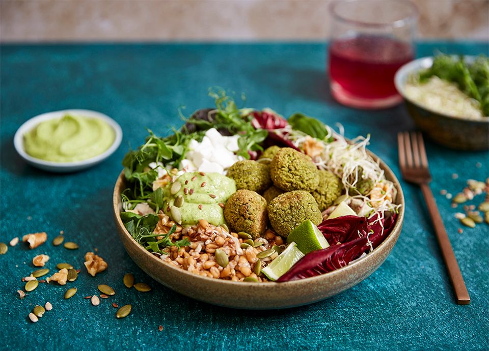 A bowl containing green meatballs, grains, rocket, radicchio and sauces against a light blue background, with seeds and nuts scattered over the top.