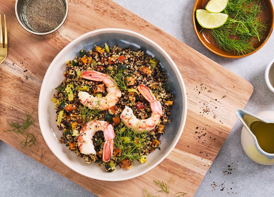 Quinoa and vegetable salad with three large prawns and fennel, served in a bowl on a wooden chopping board.