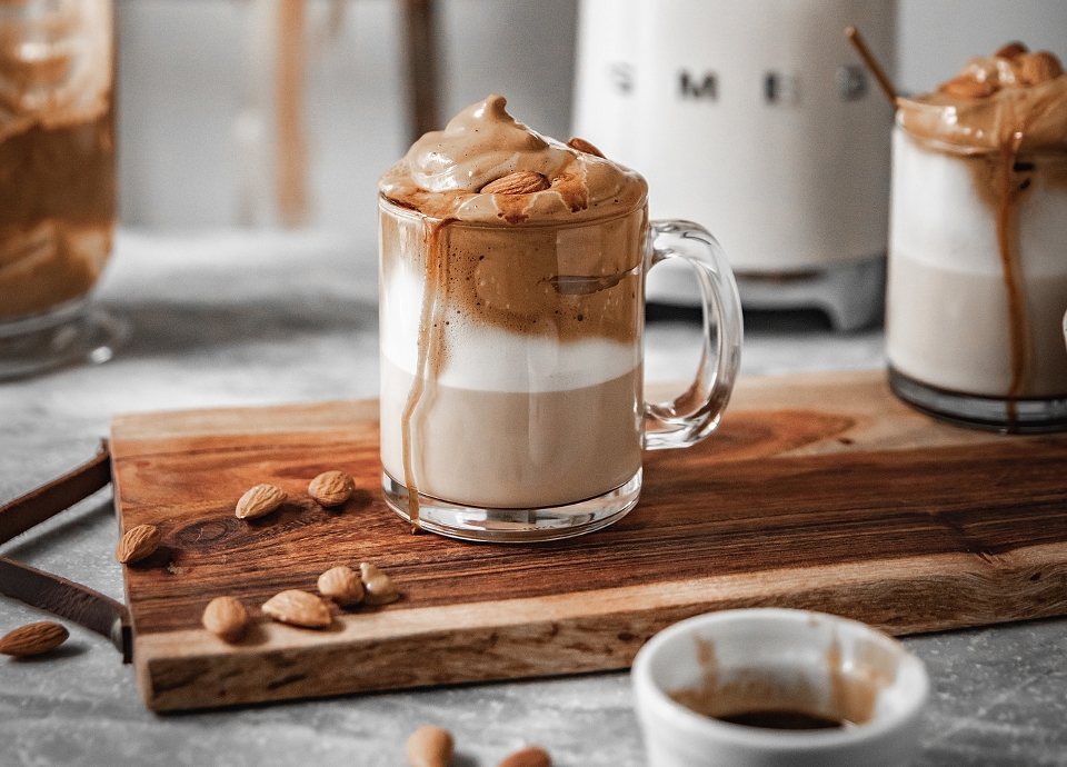 A glass filled with layers of milk and coffee, topped with a thick caramel foam and whole almonds.
