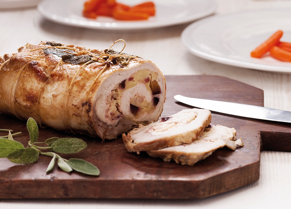 Sliced rolled roast on a chopping board with a knife and sage. In the background, white plates with baby carrots.
