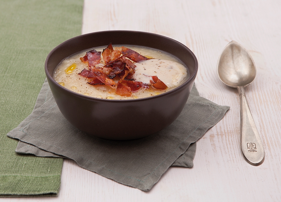 A dark bowl containing a light velouté, crispy speck and truffle cream, served with a spoon on a grey napkin.