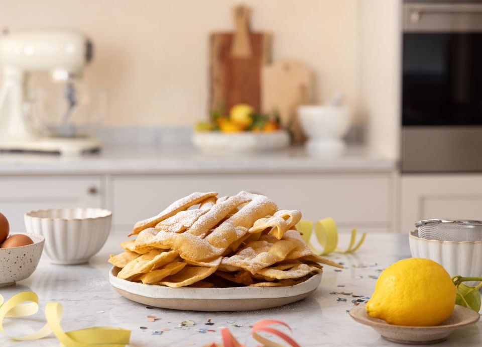 A tray of crispy chiacchiere dusted with icing sugar on a marble worktop, with a lemon and ingredients in the Smeg kitchen.