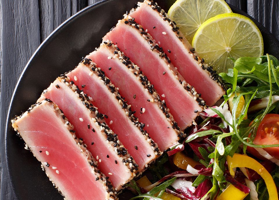 Slices of rare, pink seared tuna with a sesame crust served with a fresh garden salad and lemon on a black plate.
