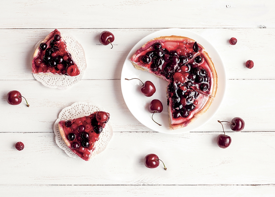 Overhead view of a berry-topped cheesecake on a white wooden table, with loose cherries and individual slices on lace doilies.