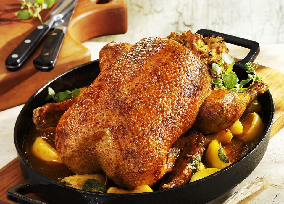 Whole golden-brown roasted duck in a black cast-iron pan, served with savory stuffing and sliced apples in a light sauce.