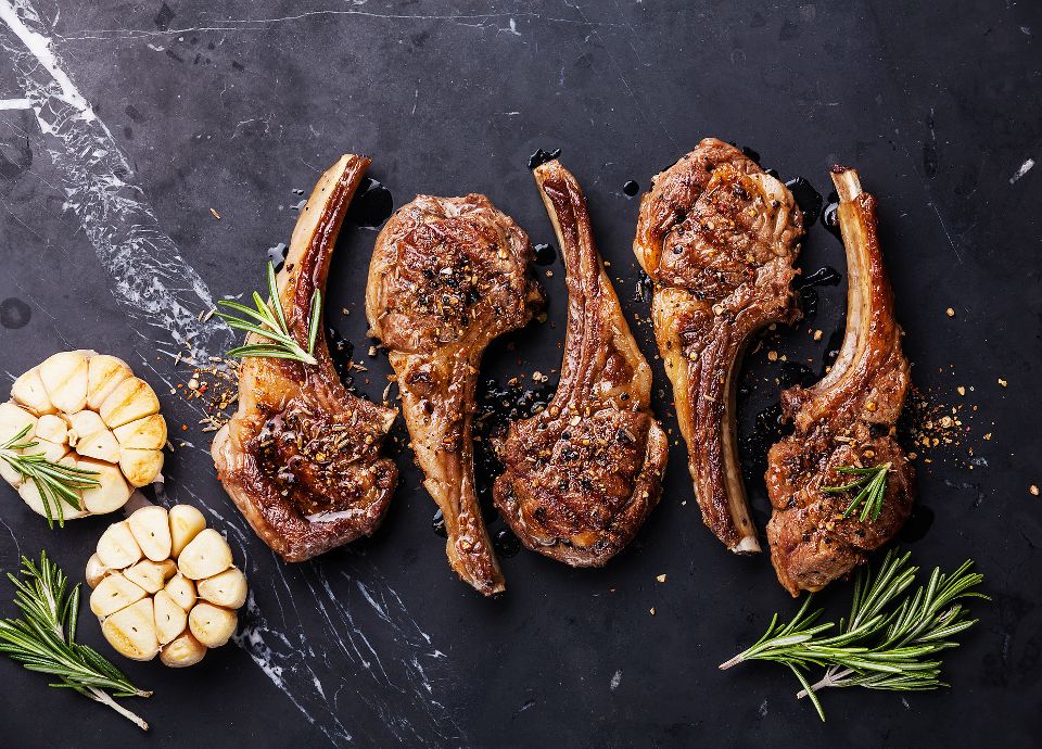 Four grilled lamb loin chops seasoned with cracked pepper and spices, garnished with rosemary on a dark marble surface.