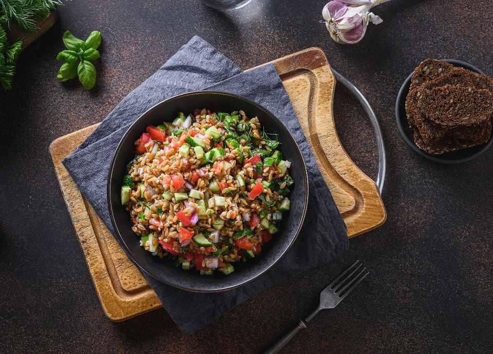 Top-down view of a dark bowl filled with spelt, diced tomatoes, cucumbers, and red onions on a rustic wooden surface.