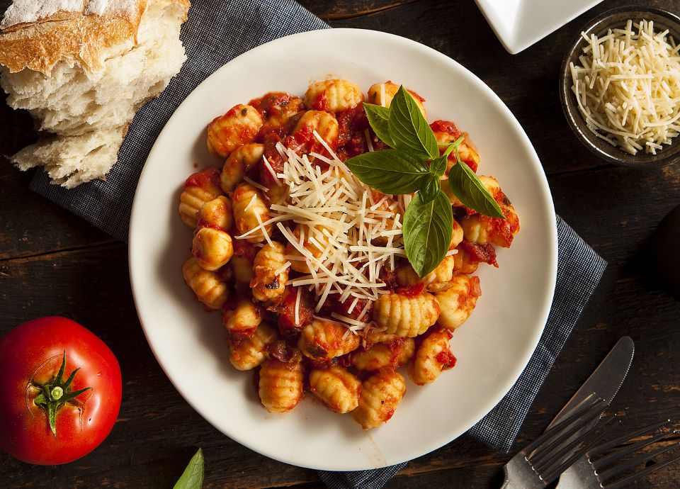 Plated potato gnocchi topped with tomato sauce, shredded cheese, and basil on a dark wood table with fresh tomatoes.