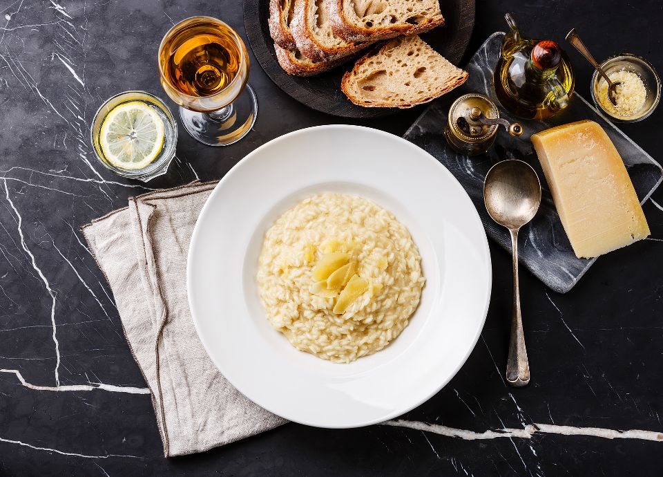 A white plate of creamy risotto topped with parmesan shavings, served with bread and a glass of white wine.