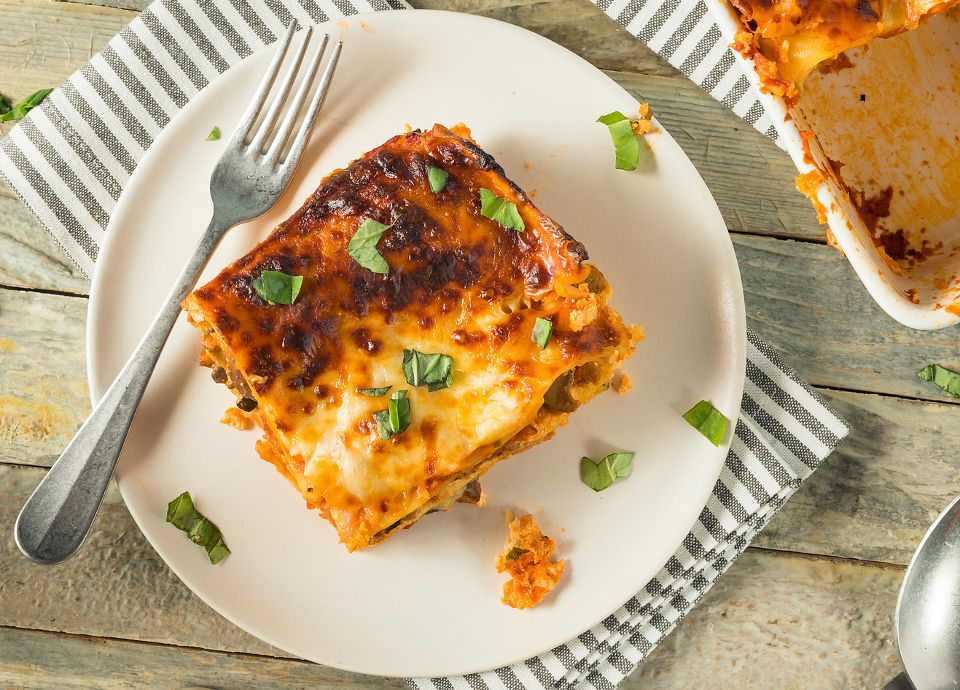Squares of lasagna with golden melted cheese and fresh basil on white plates next to a full baking dish.
