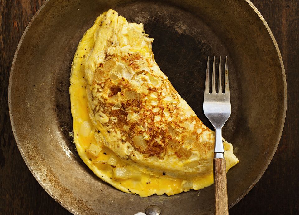 A folded golden-brown omelette with potato chunks in a rustic metal skillet, served with a wooden-handled fork.