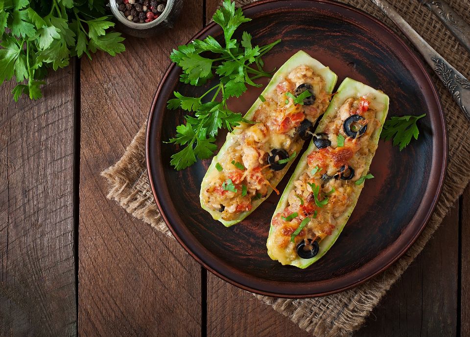 Two zucchini halves stuffed with melted cheese, tomatoes, and olives on a brown plate with fresh parsley and peppercorns.