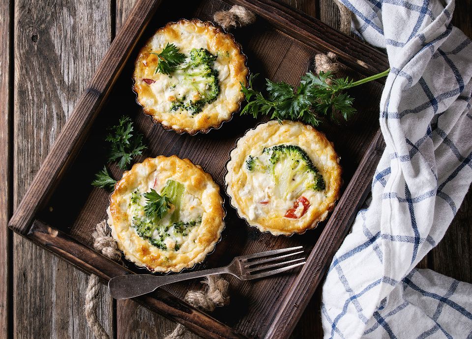 Three golden mini flans with broccoli florets in a wooden tray, garnished with fresh parsley and a silver fork nearby.