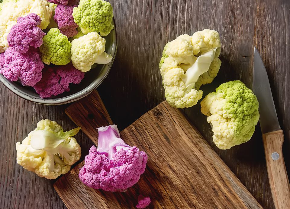 Purple, green, and white cauliflower florets on a wooden cutting board with a knife on a dark wood table.