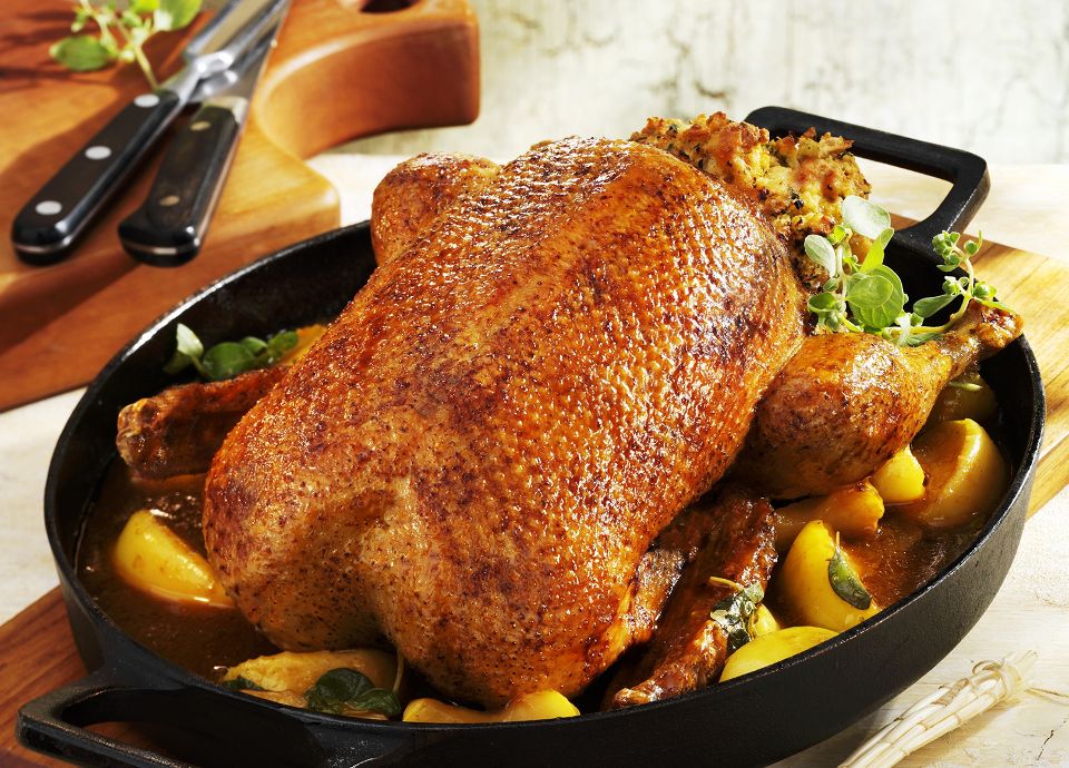 A whole golden-roasted guinea fowl in a cast iron pan, surrounded by roasted fruit and topped with herb stuffing.