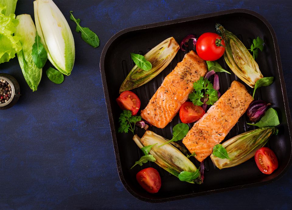 Grilled salmon fillets in a ribbed pan with charred chicory, halved cherry tomatoes, and fresh herbs.