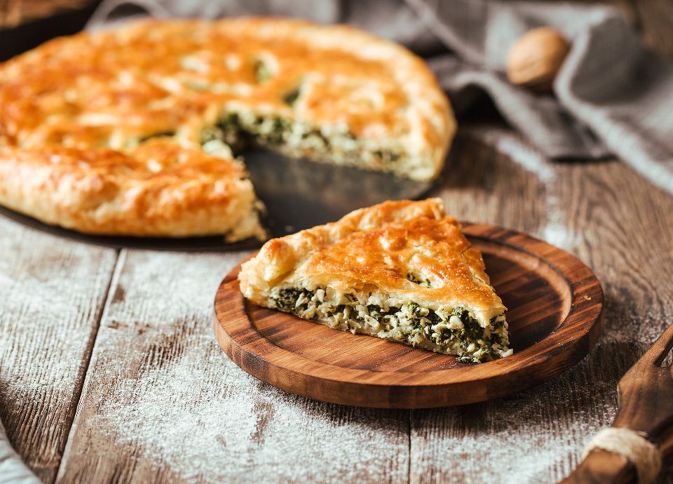 A slice of golden-brown flaky pastry filled with greens and cheese on a wood plate, with the whole pie behind it.