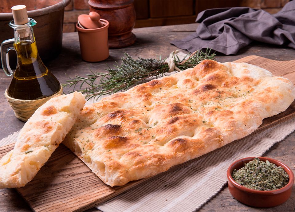 Golden-brown flatbread on a wooden board with olive oil, rosemary sprigs, and a bowl of dried herbs.