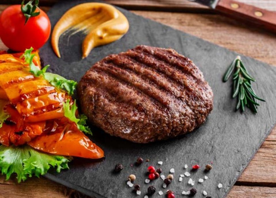 A grilled beef patty served on a slate platter with roasted peppers, fresh lettuce, a tomato, and mustard.