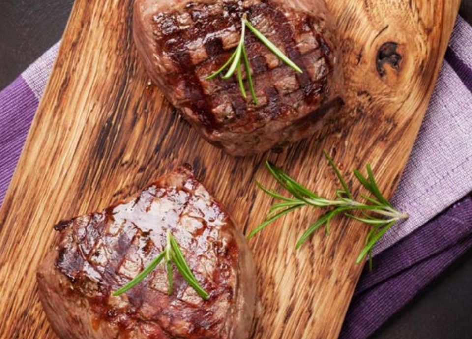 Two seared beef steaks garnished with rosemary sprigs on a wooden cutting board over a purple cloth.