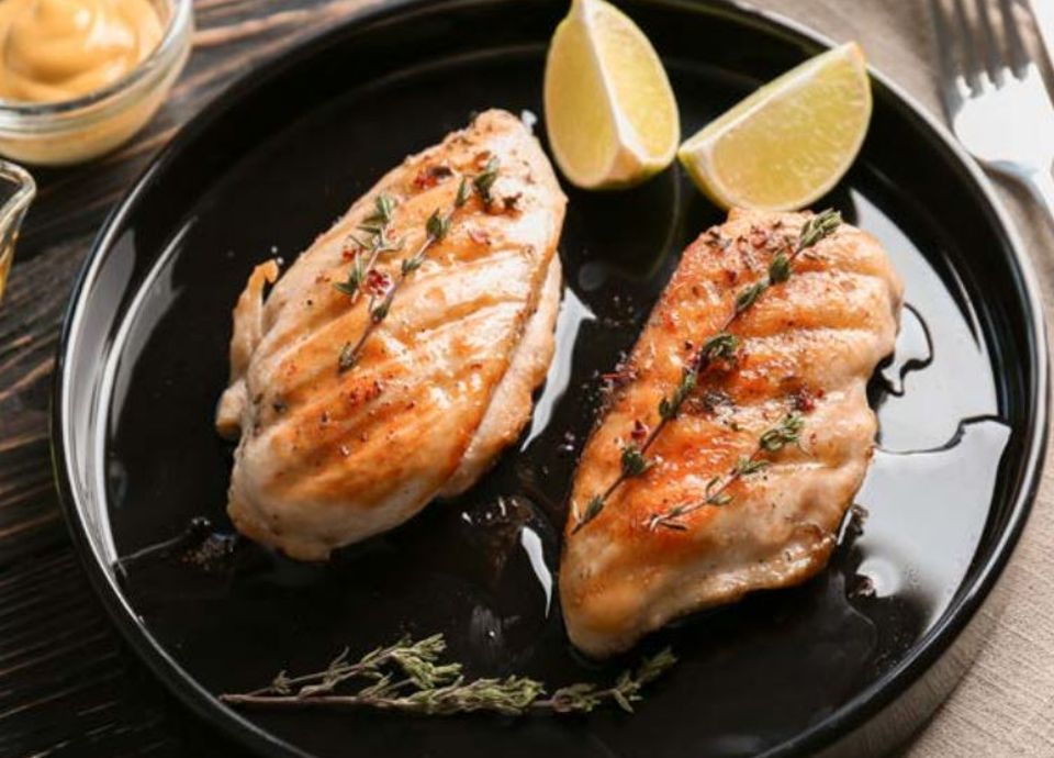 Two grilled chicken breasts garnished with thyme sprigs and lime wedges on a black plate.