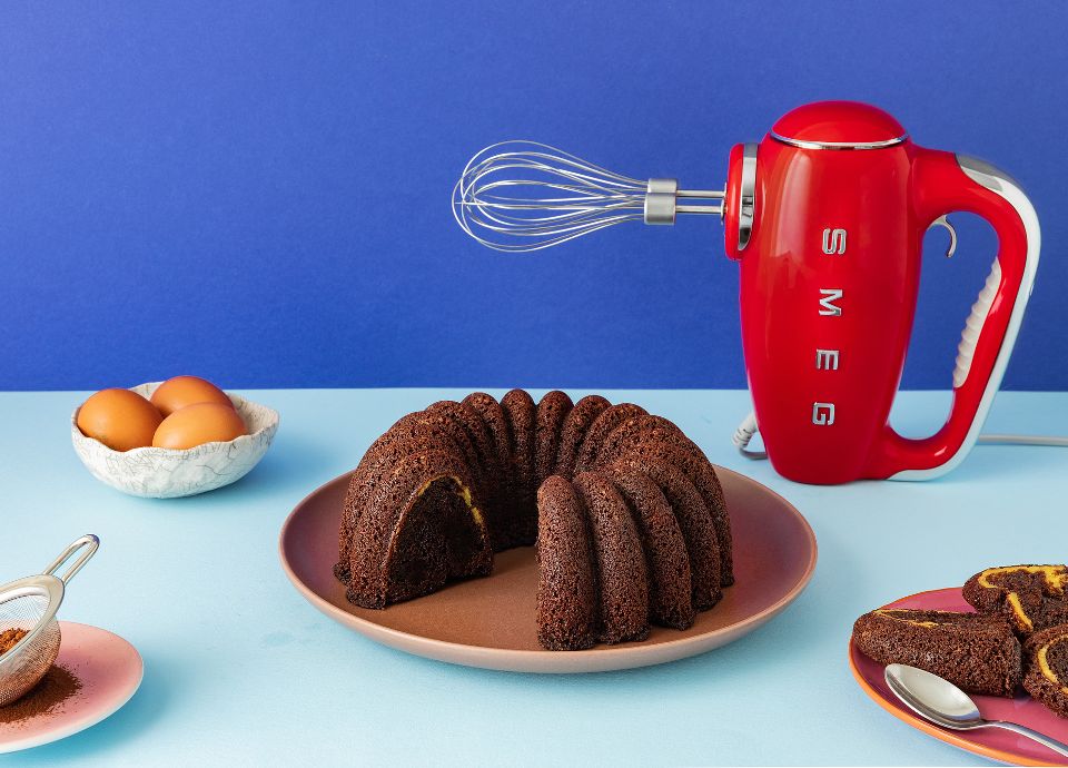 A red Smeg hand mixer with whisk attachment stands beside a sliced chocolate bundt cake on a blue background.