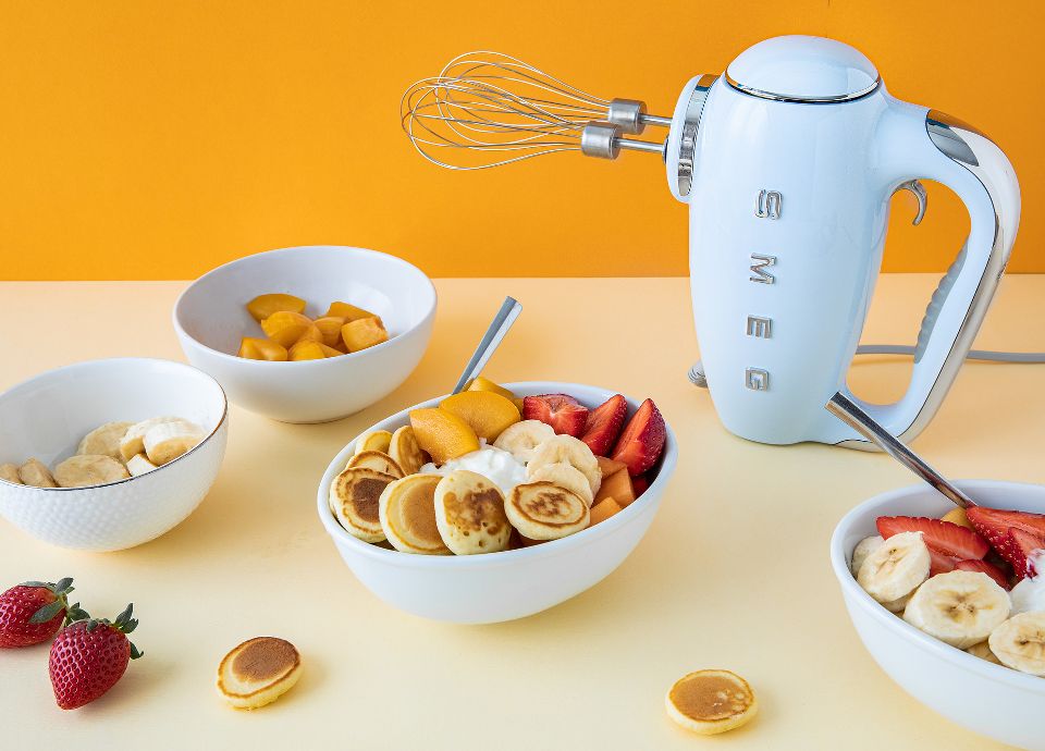 A pastel blue Smeg kettle next to bowls of mini pancakes topped with fresh strawberries, bananas, and syrup.