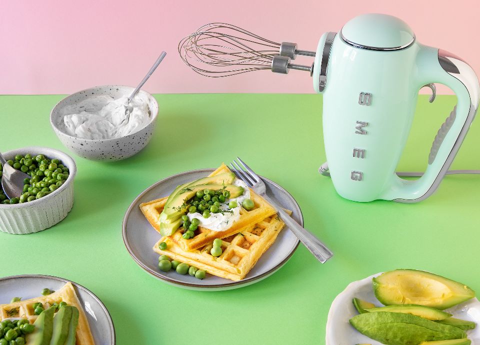 Savoury waffles topped with cream cheese and peas on green backgrounds, next to a pastel green Smeg hand mixer.