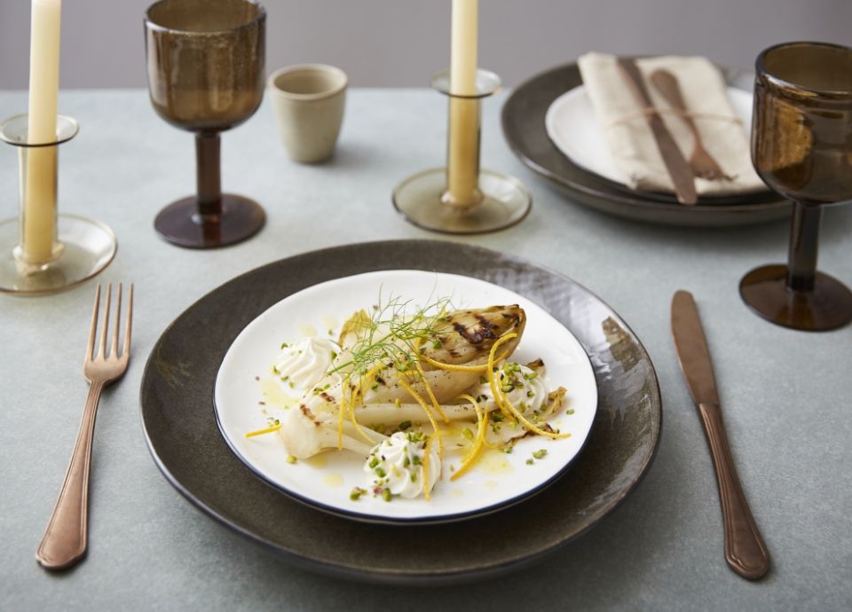 Plated grilled endive with dollops of white cheese, orange zest, and pistachios on a table with candles and glassware.