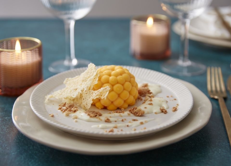 A domed pumpkin flan served with creamy parmesan sauce, crispy chips, and crushed amaretto on a white plate.