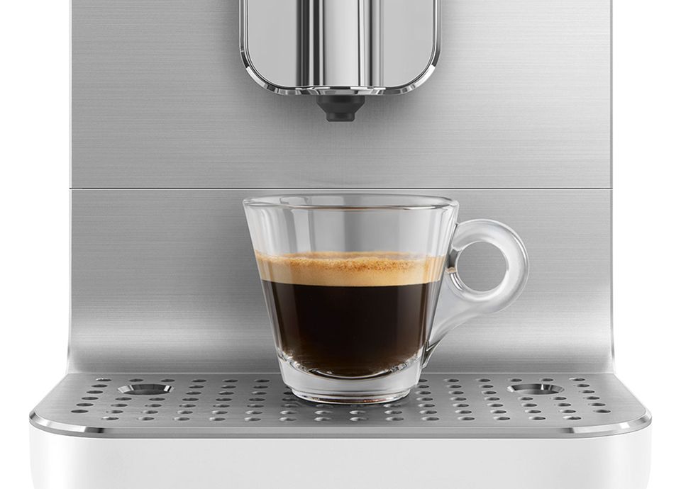 Glass cup of espresso under a stainless steel Smeg coffee machine on a perforated drip tray