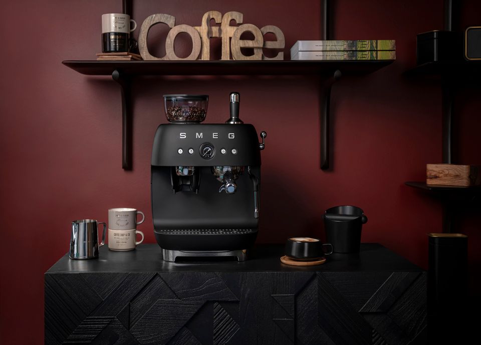 Matt black Smeg manual espresso coffee machine with built-in grinder, displayed on a dark countertop in a burgundy modern kitchen setting with cups and accessories