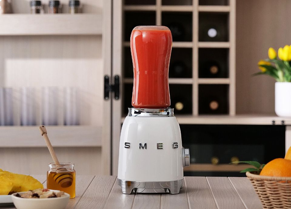 White Hand Blender with red smoothie on a kitchen counter, with honey jar, oranges, and a plant in the background.