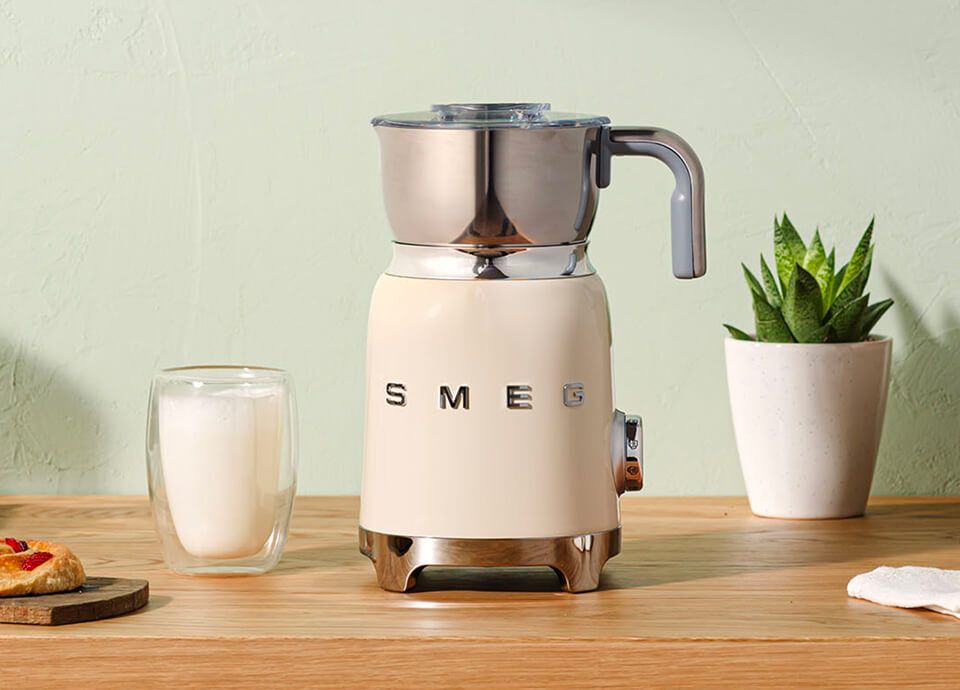 Cream SMEG milk frother with stainless steel jug on a wooden countertop, next to a glass of frothed milk and a potted plant.