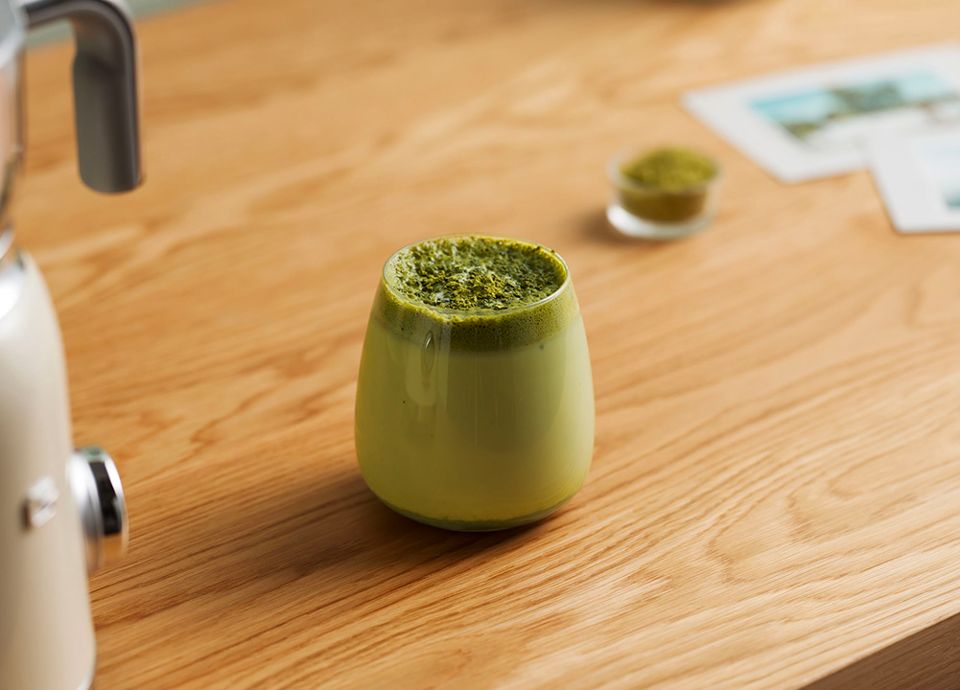 Green matcha latte in a small matte glass, topped with fine matcha powder, placed on a wooden countertop next to a kitchen appliance, with a softly blurred background.