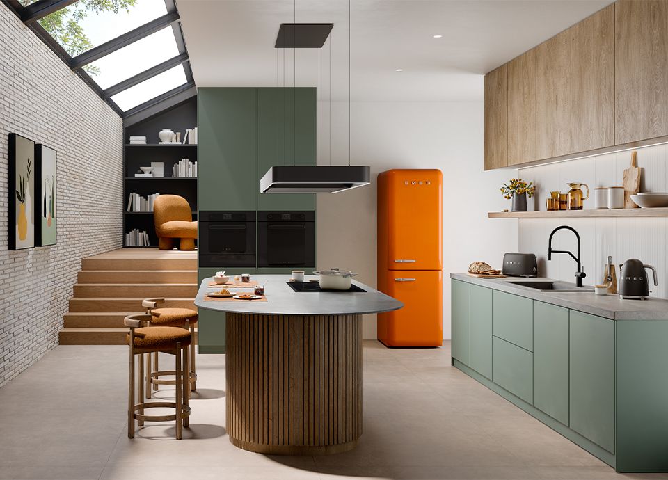 Modern kitchen with sage green cabinetry, wood finishes, a central island, SMEG ovens and suspended hood and an orange SMEG refrigerator as a standout element.