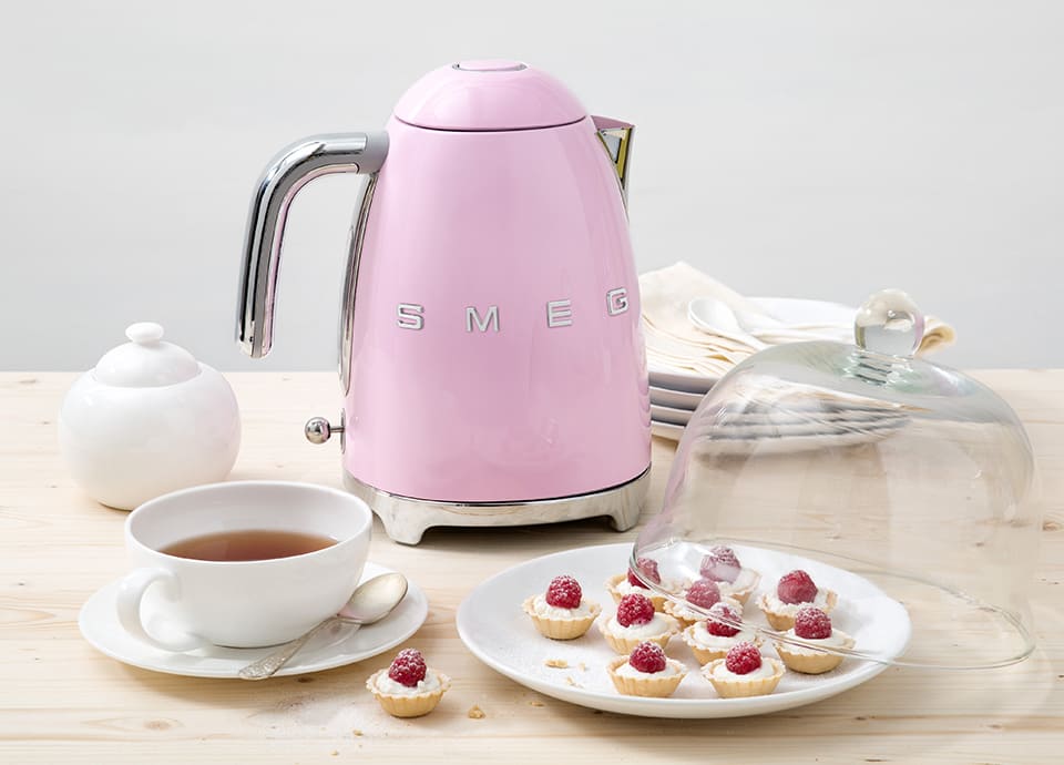 Pink electric kettle with chrome handle placed on a wooden table next to a cup of tea and small pastries under a glass dome.